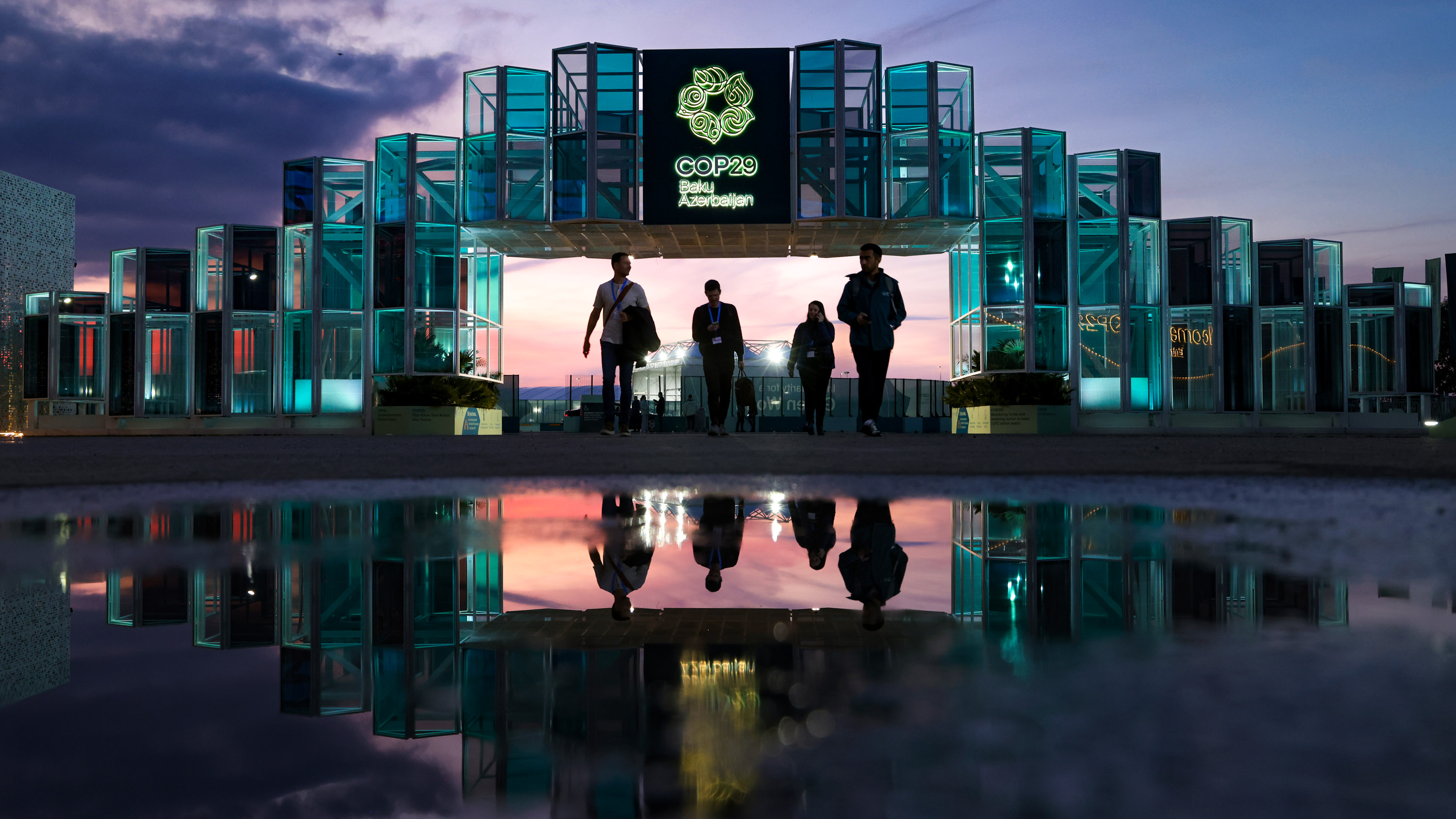 People are reflected in a puddle as they approach the illuminated gate outside the conference venue prior to the UNFCCC COP29 Climate Conference